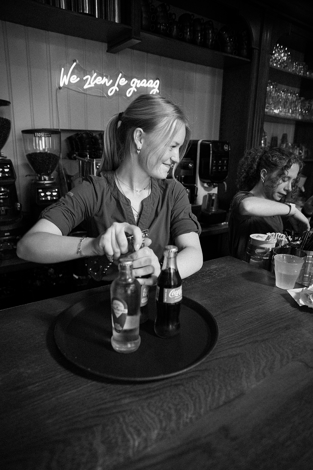 Medewerkster opent flesjes achter de bar onder het neonbord We zien je graag